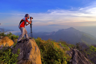 Side view of man photographing while standing on rock against sky