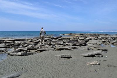 Rear view of man standing on rocks at beach against blue sky