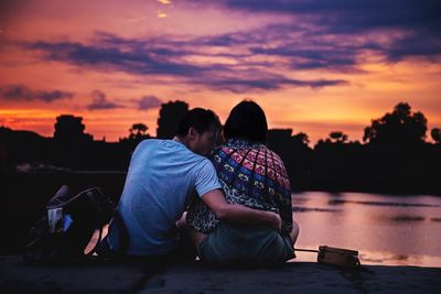 Rear view of couple sitting on shore against sky during sunset