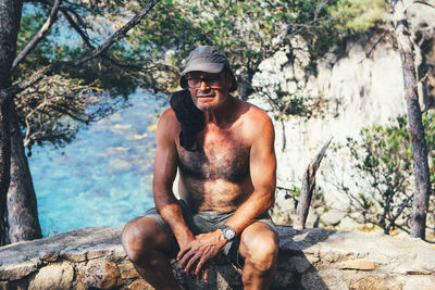 Portrait of smiling young man sitting on rock against trees