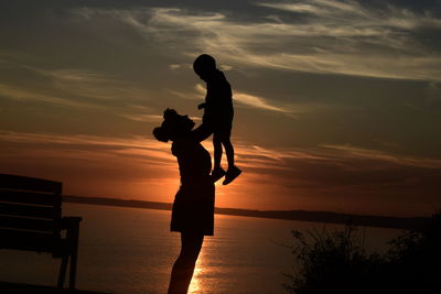 Silhouette mother lifting son while standing at beach against sky during sunset