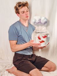 Young man holding fish tank while kneeling on bed at home