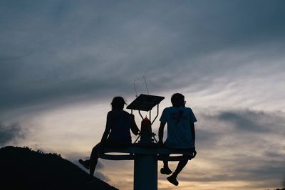 Low angle view of man and woman sitting on tower against sky during sunset