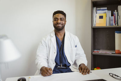 Portrait of smiling male healthcare worker wearing lab coat sitting at desk in clinic