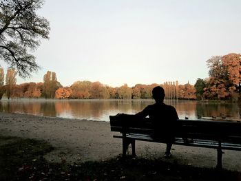 Silhouette man sitting by lake against clear sky