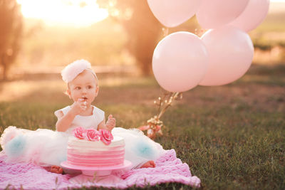 Baby girl 1 year old eating creamy birthday cake sitting on green grass with pink balloons outdoors 
