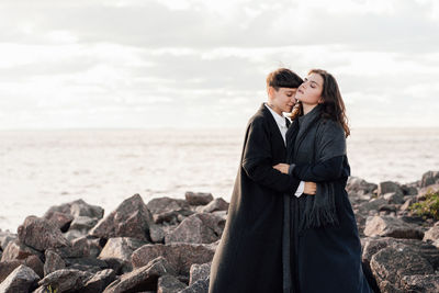 Lesbian women embracing while standing on rock against sea and sky