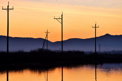 Silhouette electricity pylon by lake against romantic sky at sunset