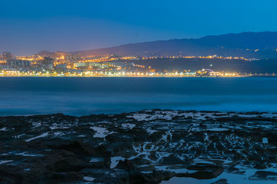 Illuminated city by sea against sky during sunset