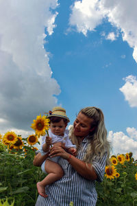 Mother holding boy with sunflower at field against sky