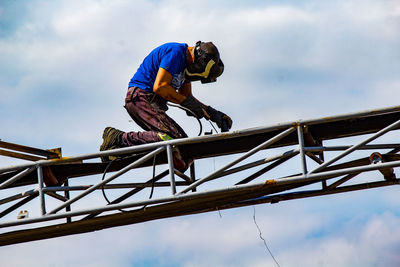 Low angle view of man working on metallic structure against sky