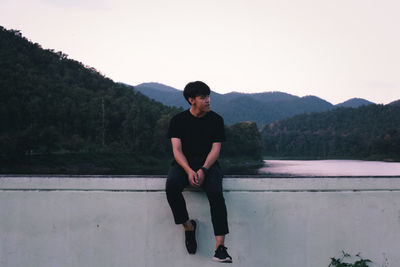 Full length of young man sitting on retaining wall against river and forest