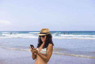 Smiling woman using mobile phone while standing at beach against sky