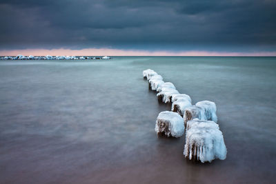 Frozen wooden posts in sea against cloudy sky