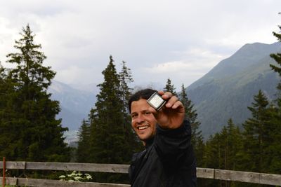 Portrait of smiling man holding digital stopwatch while standing by railing on mountain against sky