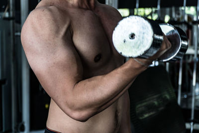 Midsection of shirtless man lifting dumbbell in gym