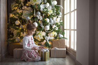 Full length of girl sitting by christmas tree at home