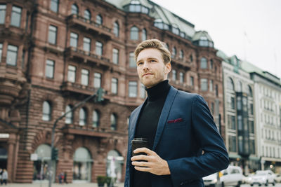 Low angle view of businessman with disposable cup looking away while standing against building in city