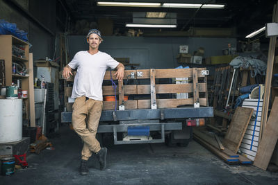Full length portrait of carpenter leaning on pick-up truck at workshop
