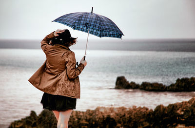 Rear view of woman holding umbrella while walking against sea at beach