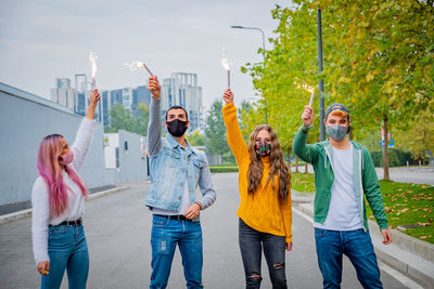 Friends wearing mask holding sparkler standing on road