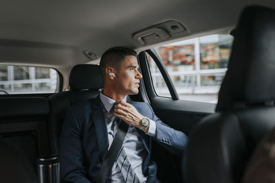 Businessman adjusting tie and looking through window while sitting in car