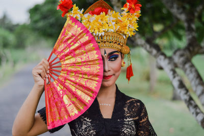 Close-up of young woman wearing headdress while holding folding fan against trees