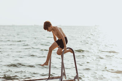 Playful shirtless boy balancing on ladder against sea during sunny day