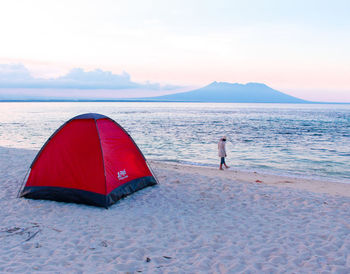 Rear view of young woman camping at beach against sky during sunset