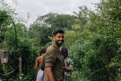 Portrait of happy man holding drink standing amidst plants in garden