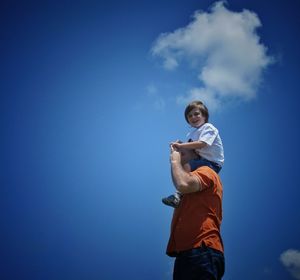 Low angle view of father carrying happy son on shoulders against blue sky