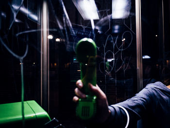 Cropped hand of woman holding telephone at booth seen through glass during night