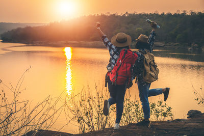 Full length of friends standing by lake against sky during sunset