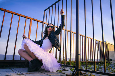 A ballerina in tutu sitting at the fence on the roof at sunset in boots and a jacket
