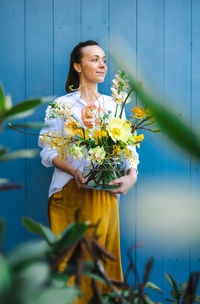 Woman in yellow skirt and white shirt with bouquet of peonies in vase against the blue background