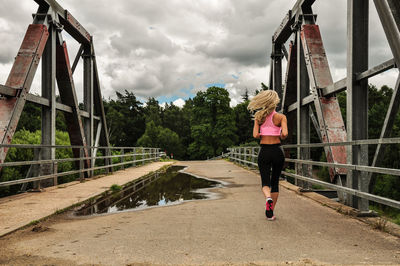 Rear view full length of woman jogging on bridge against cloudy sky