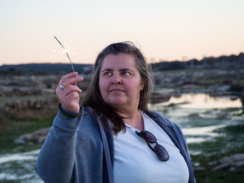 Woman holding sparkler against sky during sunset