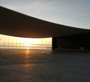 Empty road by silhouette buildings against sky during sunset