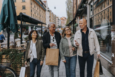 Smiling male and female senior friends holding shopping bags and talking while walking on sidewalk