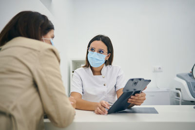 Female client in mask sitting in modern beauty clinic and discussing skin care procedures with cosmetologist during appointment