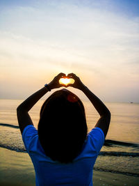 Rear view of woman making heart shape while standing at beach against sky during sunset