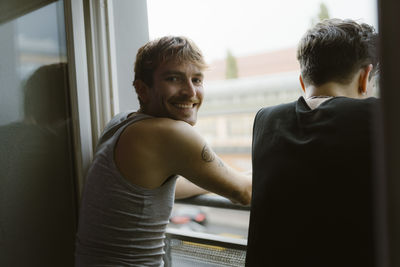 Smiling gay man looking over shoulder while standing with friend near window in apartment