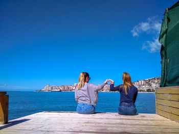 Rear view of female friends making heart shape on pier in sea against blue sky