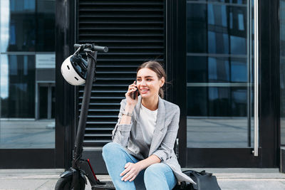 Young businesswoman sitting on an electric push scooter near a building and talking on mobile phone	