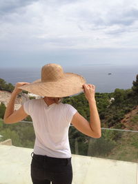 Rear view of woman holding straw hat while looking at sea