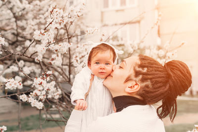 Mother kissing daughter while standing against tree outdoors