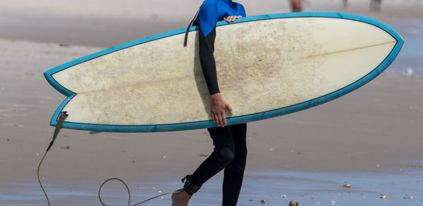 Side view of a surfer wearing a black and blue wetsuit carrying his surfboard into the ocean.