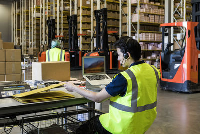 Rear view of confident young male customer service representative looking at package while sitting with laptop in wareho