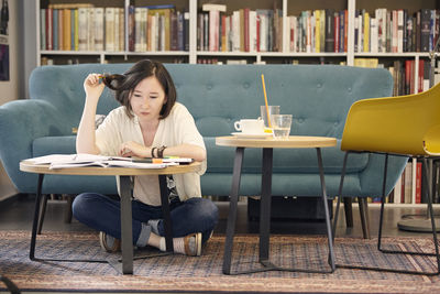 Full length of young woman studying while sitting by sofa on carpet at home