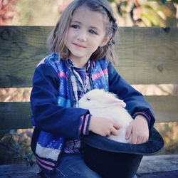 Close-up of smiling girl holding rabbit in hat while sitting on bench at park